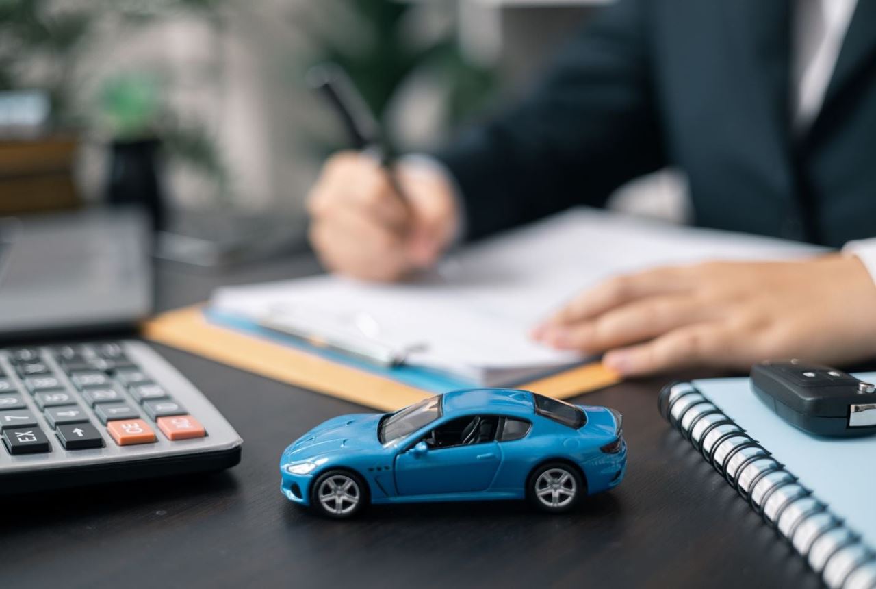 Ruka koja piše po dokumentu na radnom stolu. U prvom planu maketa plavog automobila.
