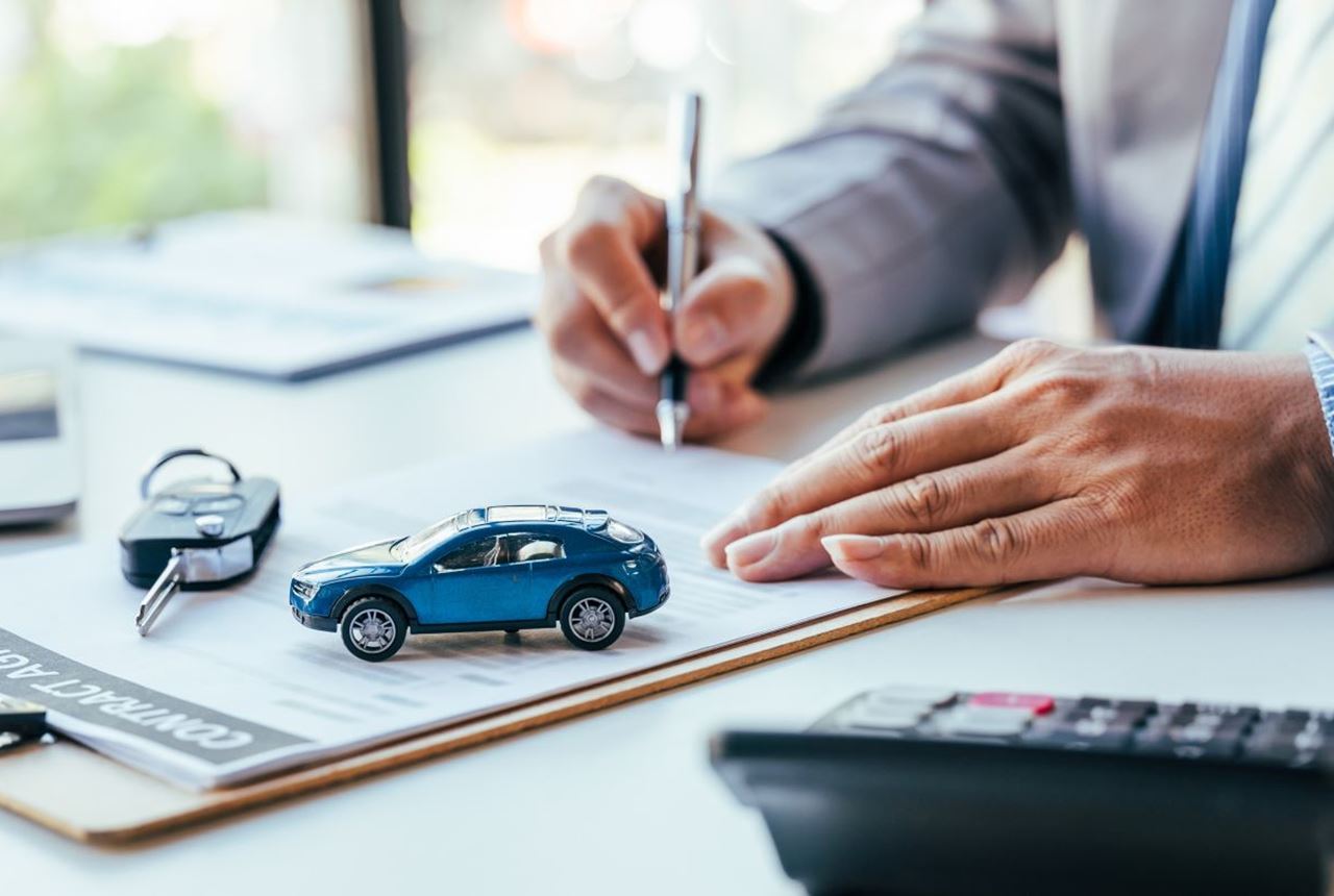Ruka poslovnog čoveka potpisuje dokument na radnom stolu. U prvom planu je maketa plavog automobila.